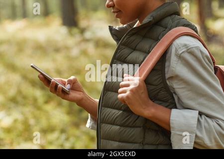 Beschnittene Seitenansicht einer jungen afroamerikanischen Frau, die mit dem Smartphone unterwegs ist, während sie im Wald wandern und den Weltraum kopieren kann Stockfoto