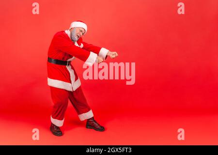 In voller Länge hart arbeitenden älteren Mann mit grauem Bart trägt weihnachtsmann Kostüm stehen mit Faust oder Zug Geste, um unsichtbare Seil zu ziehen. Innenaufnahme des Studios isoliert auf rotem Hintergrund. Stockfoto