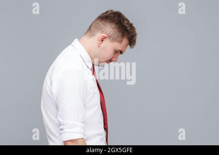 Seitenansicht Profil Porträt von jungen schönen traurigen Mann in weißem Hemd und Krawatte stehen mit Kopf nach unten und traurig Gesicht suchen. Indoor isoliert auf grauem Hintergrund. Stockfoto