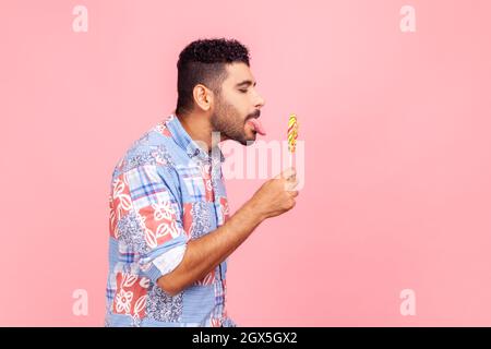 Seitenansicht des bärtig schönen Mannes mit blauem Casual Shirt Stehen schmeckt Lollipop, träumen lecken um süße zuckerhaltige Regenbogenbonbon. Innenaufnahme des Studios isoliert auf rosa Hintergrund. Stockfoto