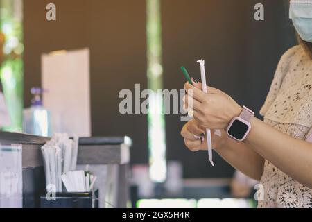 Hände einer Frau, die eine Operationsmaske mit zwei Strohhalmen trägt, die sich für den Eiskaffee aus einem Café vorbereiten. Eine Frau, die grünes Stroh auspackelt, eingewickelt Stockfoto