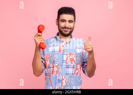 Porträt eines jungen erwachsenen attraktiven Mannes mit dunklem Haar und Bart, der den Daumen hochzeigt und das rote Festnetztelefon hält, guter Service im Call Center. Innenaufnahme des Studios isoliert auf rosa Hintergrund. Stockfoto
