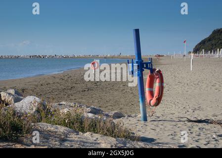 Blick auf leeren Strand - roter Rettungsring auf blauem Pfahl am Sandstrand in der Nähe des Meerwassers, Steine von Klippen am Horizont und klaren Himmel, zwischen Ligurien/Toscana Stockfoto