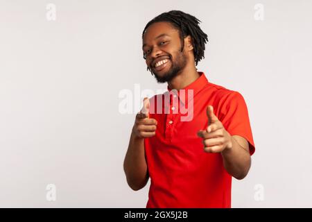 Porträt eines Mannes mit Dreadlocks in einem roten T-Shirt im lässigen Stil, der spielerisch mit den Fingerpistolen auf die Kamera zeigt und flirtet und den glücklichen Gewinner wählt. Innenaufnahme des Studios isoliert auf grauem Hintergrund. Stockfoto