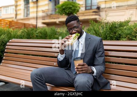 Mittagspause. Junger afroamerikanischer Geschäftsmann, der Sandwich und Kaffee genießt und auf einer Bank in der Nähe des Bürogebäudes sitzt Stockfoto