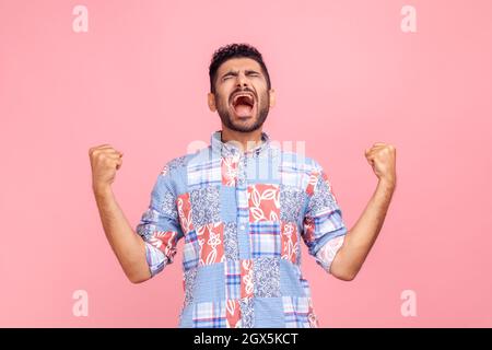 Porträt eines überglücklich aussehenden Mannes mit Bart im legeren Hemd, der mit erhobenen Fäusten und Schreien eine siegreiche Geste zum Ausdruck bringt und den Sieg feiert. Innenaufnahme des Studios isoliert auf rosa Hintergrund. Stockfoto