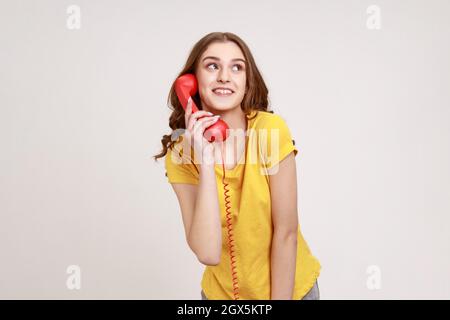 Positive glückliche junge Frau mit braunem Haar in gelbem T-Shirt sprechendes Festnetztelefon, das in der Hand hält und mit verträumtem Blick wegschaut. Innenaufnahme des Studios isoliert auf grauem Hintergrund. Stockfoto