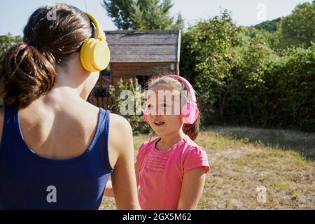 Zwei Mädchen schauen sich gegenseitig an, während sie gelbe und rosa Kopfhörer im Hinterhof tragen. Zwei Mädchen, die über Kopfhörer Musik hören, sehen sich gegenseitig aus. Stockfoto
