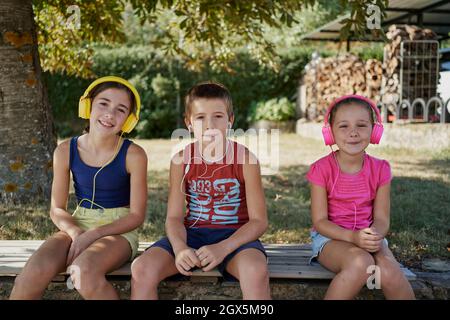 Drei Kinder lächeln vor der Kamera, während sie über s Steinmauer sitzen und Musik hören. Drei Kinder verwenden ihre Kopfhörer und Kopfhörer für LIS Stockfoto