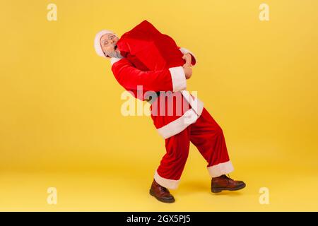 In voller Länge ältere Mann mit grauem Bart trägt weihnachtsmann Kostüm trägt große schwere Tasche mit Weihnachtsgeschenken, gratuliert zum neuen Jahr. Innenaufnahme des Studios isoliert auf gelbem Hintergrund. Stockfoto