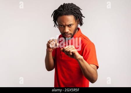 Aggressiver bärtiger Mann mit Dreadlocks, der ein rotes T-Shirt im lässigen Stil trägt, geballte Fäuste zum Boxen bereithält, Kampfkunsttrainer, Selbstverteidigung. Innenaufnahme des Studios isoliert auf grauem Hintergrund. Stockfoto