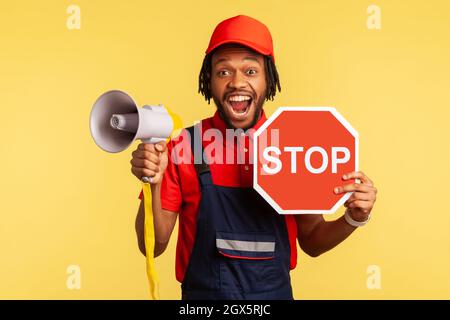 Porträt eines aufgeregten bärtigen Mannes mit blauen Overalls und rotem T-Shirt, der die Kamera anschaut, das rote Stoppschild und das Megaphon in den Händen hält. Innenaufnahme des Studios isoliert auf gelbem Hintergrund. Stockfoto