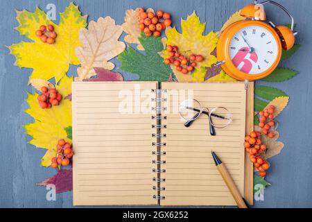 Orangefarbener Wecker, ledergefütterter Notizblock, Stift und Gläser auf einem Hintergrund mit trockenen, farbenfrohen Herbstblättern. Konzept für die Planung des Arbeitstages. Herbst tim Stockfoto