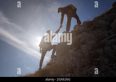 Hilfe- und Hilfekonzept. Silhouetten von zwei Menschen, die Berge besteigen und gegen den blauen Himmel helfen. Gehen Sie in den Wolken. Discovery Travel Direc Stockfoto