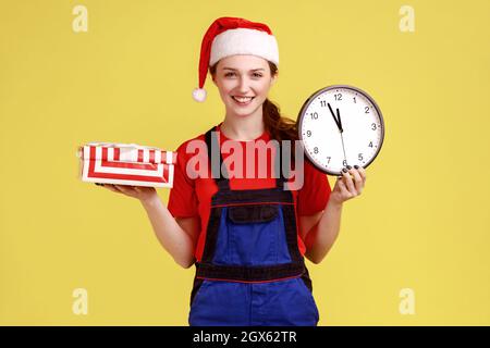 Lächelnde Kurierin mit eingepackter Geschenk-Box und Wanduhr, schnellem Lieferservice in der Zeit, mit blauen Overalls und weihnachtsmannhut. Innenaufnahme des Studios isoliert auf gelbem Hintergrund. Stockfoto