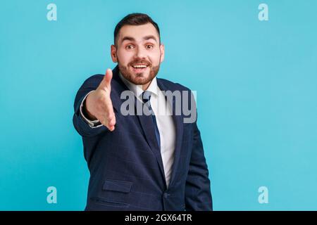 Porträt eines freundlichen, positiv schönen Mannes mit Bart, der einen dunklen Anzug im offiziellen Stil trägt, der Hand zum Händeschütteln gibt, einladend ist und sich kennenlernt. Innenaufnahme des Studios isoliert auf blauem Hintergrund. Stockfoto