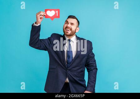 Optimistischer bärtiger Mann im offiziellen Anzug, hält das Herz des sozialen Netzwerks wie ein Symbol über den Kopf, schaut auf das Symbol und zwinkert. Innenaufnahme des Studios isoliert auf blauem Hintergrund. Stockfoto