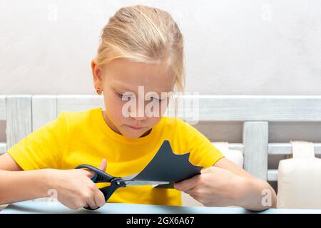 Ein Mädchen schneidet zu Hause mit eigenen Händen eine Halloween-Fledermaus aus schwarzem Bastelpapier mit einer Schere. Startseite Halloween Urlaubskonzept. Stockfoto