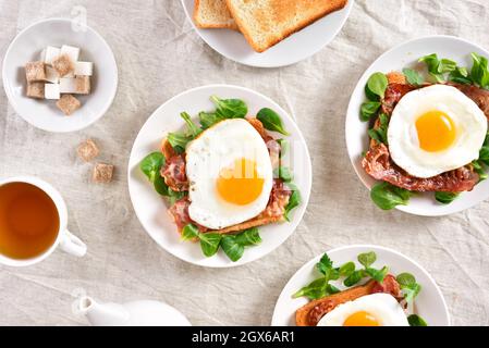 Offene Sandwiches mit Spiegeleiern, Speck und Gemüseblättern auf dem Teller auf hellem Hintergrund. Draufsicht, flach liegend Stockfoto