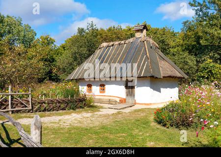 Rekonstruktion eines alten traditionellen ukrainischen Landhauses mit Reetdach vor dem Hintergrund eines Sommergartens mit Blumenbeet. Stockfoto