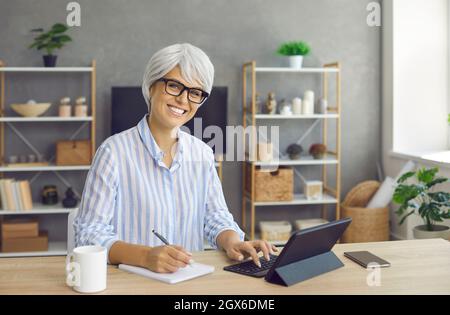 Ältere Geschäftsfrau, die am Schreibtisch sitzt, einen Tablet-Computer benutzt und in einem Notebook schreibt Stockfoto