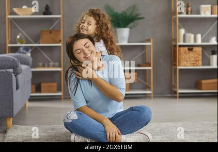 Die kleine Tochter umarmt ihre fröhliche Mutter, die im Wohnzimmer auf dem Boden sitzt. Stockfoto