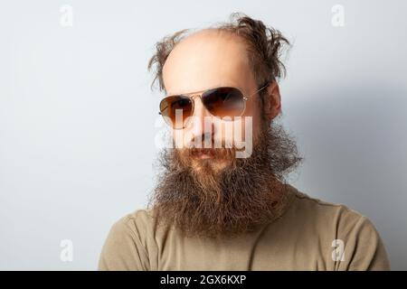 Porträt eines erwachsenen, gutaussehenden, ernsthaften Mannes, der mit ruhigem Gesichtsausdruck auf die Kamera schaut, eines kahlen bärtigen Mannes mit T-Shirt und Sonnenbrille. Innenaufnahme des Studios isoliert auf grauem Hintergrund. Stockfoto