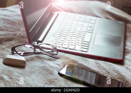 Ein Laptop, eine Brille, Kopfhörer und ein Telefon liegen auf dem Bett, während sie aus der Ferne arbeiten. Stockfoto