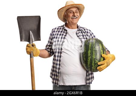 Lächelnder, reifer Bauer, der eine Schaufel und eine Wassermelone auf weißem Hintergrund hält Stockfoto