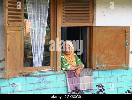 Eine ältere muslimische Frau, die einen grünen Hijab trägt und aus ihrem Fenster in der Nähe der Stadt Bukittinggi auf West-Sumatra, Indonesien, blickt. Stockfoto