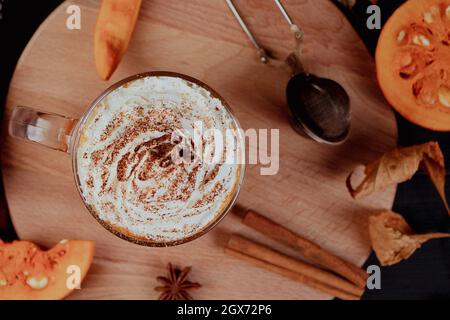 Kürbis-Kaffee-Latte mit Schlagsahne auf dunklem Hintergrund. Heißer Herbsttrunk mit Zimt und Gewürzen im Glaskrug auf Holztablett. Herbst und Gemütlichkeit c Stockfoto