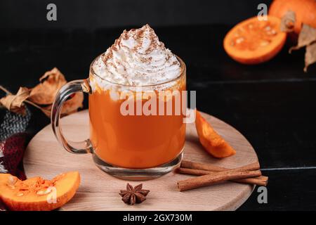 Kürbis-Kaffee-Latte mit Schlagsahne auf dunklem Hintergrund. Heißer Herbsttrunk mit Zimt und Gewürzen im Glaskrug auf Holztablett. Herbst und Gemütlichkeit c Stockfoto