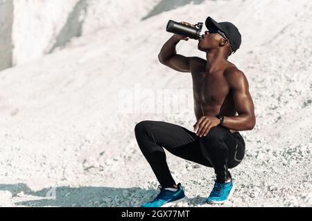 Fitness-Athlet, afrikanischer Muskelmann, trinkt Wasser in einer wiederverwendbaren Flasche, Mann mit Wasser nach dem Training an einem sonnigen Tag in der Natur, Sportkonzept, Motiv Stockfoto