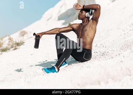 Erschöpfter sportlicher afroamerikanischer Mann, muskulöser Mann in Sportkleidung, mit Kopfhörern, mit einer Flasche Trinkwasser, Sport im Freien in der absichtserklärung Stockfoto