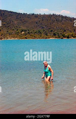 Lächelnde ältere Frau im Badeanzug, die kniehoch im See mit Hilfe einer Krücke steht. Stockfoto