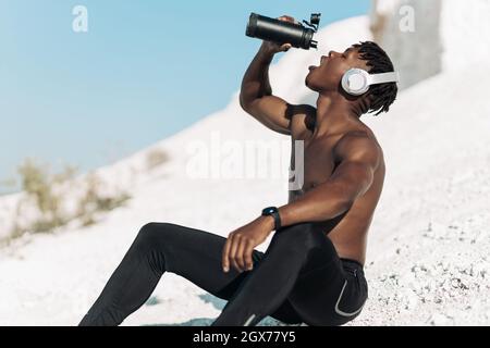 Fitness-Athlet, afrikanischer Muskelmann, trinkt Wasser in einer wiederverwendbaren Flasche, Mann mit Wasser nach dem Training an einem sonnigen Tag in der Natur, Sportkonzept, Motiv Stockfoto