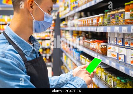 Seitenansicht Foto eines Ladenarbeiters in einer medizinischen Maske mit Schürze in schwarzer Farbe, der das Smartphone hält und die Inventarisierung der Produkte vorführt Stockfoto