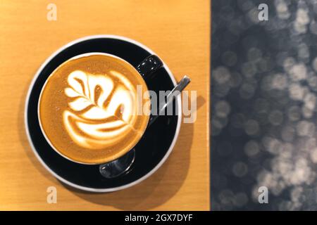 Latte Art in schöner, glänzend schwarzer Kaffeetasse auf Holztisch. Koffein Cappuccino-Getränk aus Pflanzenmilch mit Schaum. Bokeh-Hintergrund, Textraum Stockfoto