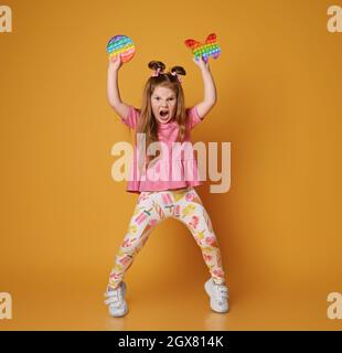 Verwöhnte, schreiende laut Kind Mädchen in bunten Kleidern gehen, um zwei sensorische Regenbogenfarben Spielzeug werfen - rund Pop it Stockfoto