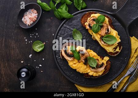 Rührei mit gebratenen Pilzen und Basilikum auf Brot auf schwarzem Tischhintergrund. Hausgemachtes Frühstück oder Brunch-Menü - Rührei und Pilze san Stockfoto