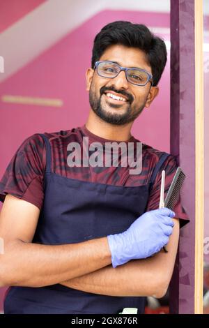 Fröhlicher Barbier im Salon, der selbstbewusst vor der Tür auf die Kamera blickt - ein Konzept von Glück, erfolgreichen Geschäften und positiven Emotionen. Stockfoto