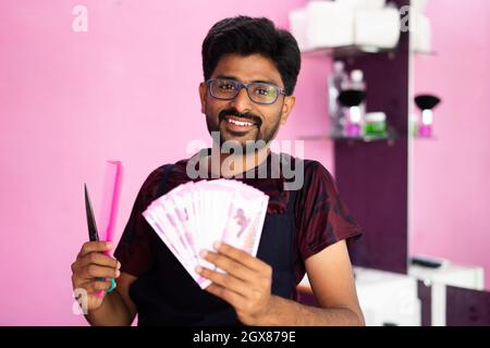 Glücklich lächelnder indischer Barbier mit Geldscheinen - Konzept des erfolgreichen Geschäfts, der Gewinngestaltung, des Bankwesens und der Finanzen. Stockfoto