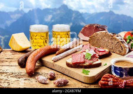 Verschiedene, trocken geräucherte Würstchen, serviert auf Schneidebrett mit Brot auf einem Holztisch und einem Becher Bier auf Berggrund Stockfoto