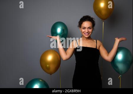 Charmante Frau in schwarzem Samtkleid, lächelt mit den Handflächen nach oben auf die Kamera und hält einen imaginären Platz für Werbung. Geburtsh Stockfoto
