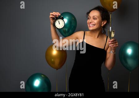 Wunderschöne elegante Frau in schwarzem Abendkleid isoliert auf grauem Hintergrund mit goldgrünen Ballons, hält eine Flöte Sekt und schaut zu Stockfoto