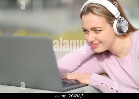 Zufriedener Teenager mit kabellosen Kopfhörern, der seinen Laptop in einem Park überprüft Stockfoto