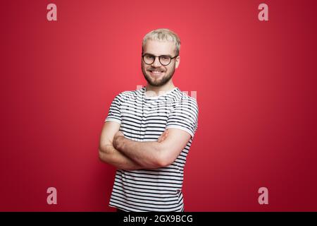 Trendy Mann mit blonden Haaren posiert an der roten Wand mit. Schöner Mann. Lächelnder Mann. Stockfoto