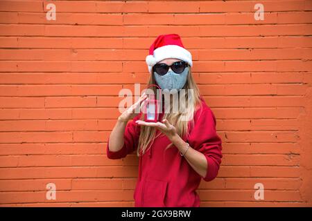 Blonde Mädchen in silberner Maske, roten Hut und schwarze Sonnenbrille hält in den Händen Weihnachtslicht. Kaukasische Frau in Weihnachtskostüm und covid Schutz Stockfoto