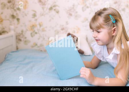 Ein kleines Mädchen leet auf dem Bett im stilvollen Schlafzimmer und liest ein blaues Buch und macht Hausaufgaben. Bildung, Home Schooling Konzept Stockfoto