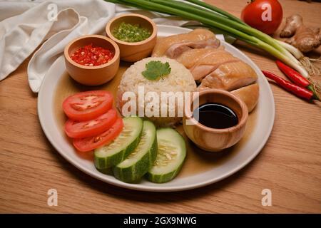 Leckerer Hainanese-Hühnerreis mit Chilisaucen, süßer Sojabohnensauce und frischem Gemüse auf einem Holztisch. Asiatisches Gourmet-Konzept. Nahaufnahme Stockfoto
